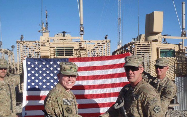 Four U.S. military personnel in MultiCam/OCP camouflage uniforms are posing in front of an American flag with armored military vehicles (likely MRAPs) visible in the background. Two of the soldiers in the foreground are shaking hands while holding rifles, suggesting a formal or ceremonial moment. The setting appears to be a forward operating base in an arid environment.