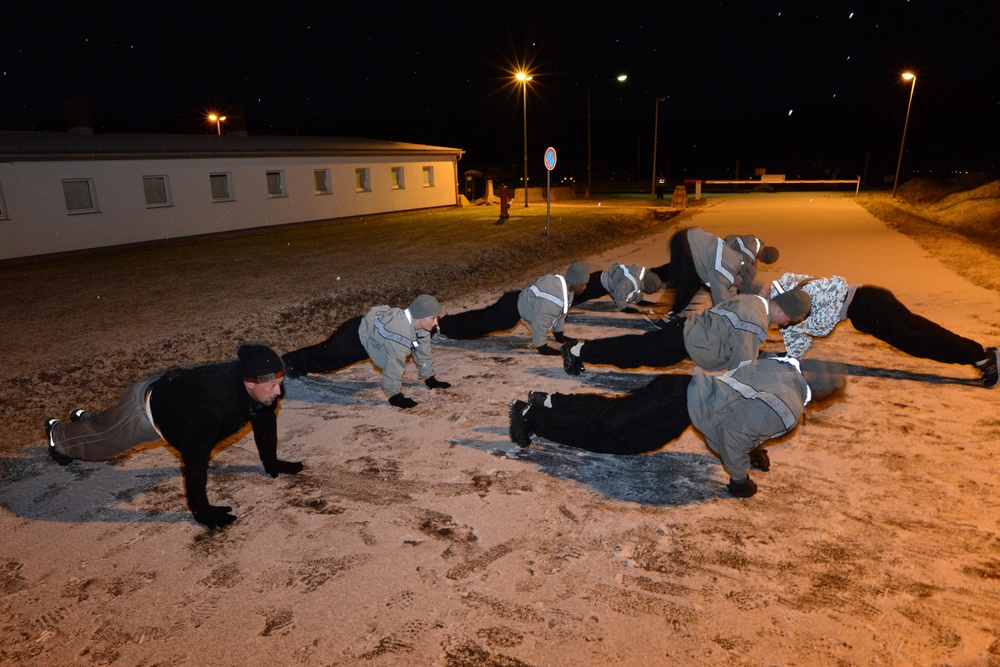 A group of individuals in dark athletic/military-style clothing with reflective stripes are performing push-ups on a snow-dusted ground surface at night. The exercise appears to be an outdoor physical training session conducted in cold winter conditions under artificial street lighting. A low-rise building and multiple street lights are visible in the background.