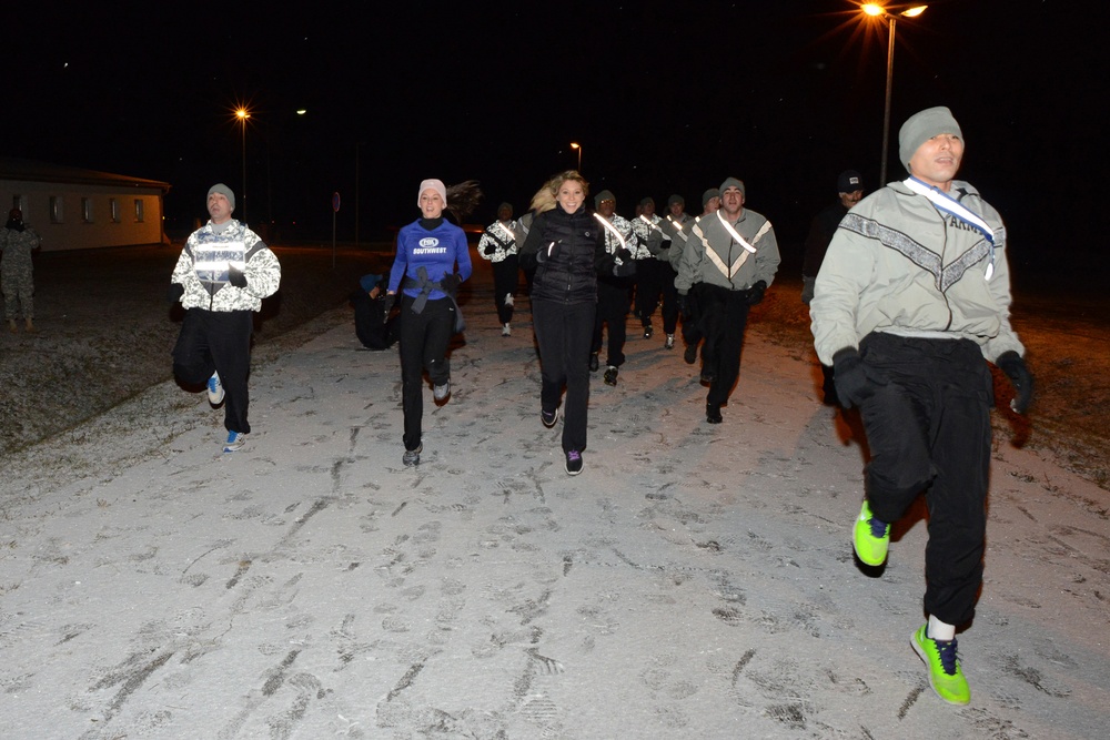 A group of people are running outdoors at night on a snow-dusted surface, likely a military base given the Army-branded clothing visible on at least one participant. The runners are dressed in cold-weather athletic gear including hats, gloves, and jackets. Street lights illuminate the scene, suggesting an early morning or nighttime physical training exercise.