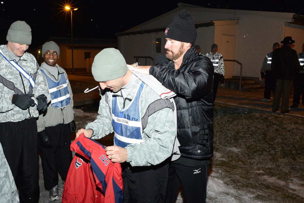 A nighttime outdoor scene at what appears to be a military installation, showing military personnel in camouflage uniforms interacting with a civilian man in a black jacket. The civilian appears to be signing or presenting a red Boston Red Sox jacket to a soldier identified by a vest label as 'SSG CELON'. The scene suggests a celebrity or notable visitor meeting with troops.