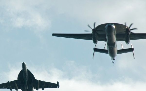 Two military aircraft are photographed from below against an overcast sky. The formation consists of a twin-engine turboprop aircraft and a single jet fighter flying in close proximity. Both aircraft are viewed from a rear/underside angle, silhouetted against light cloud cover.