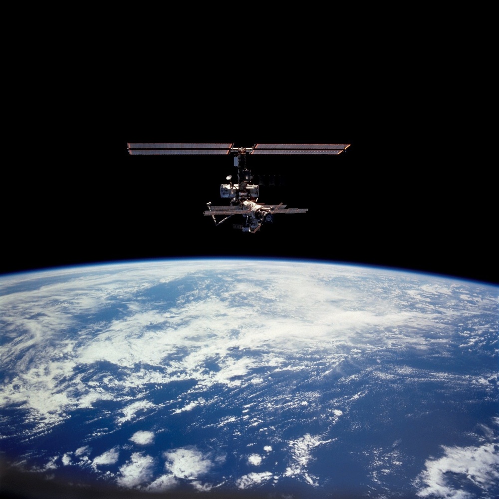A space station is photographed against the blackness of space with Earth's curved horizon visible in the lower portion of the image. The station displays a cross-shaped configuration with large solar panel arrays extending horizontally. Earth's surface below shows blue oceans, white cloud formations, and the thin luminous atmospheric limb.