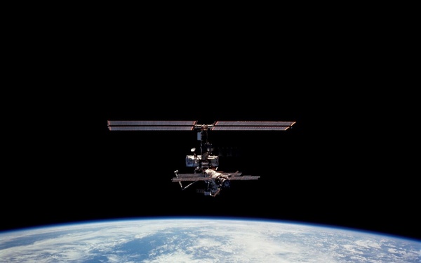A space station is photographed against the blackness of space with Earth's curved horizon visible in the lower portion of the image. The station displays a cross-shaped configuration with large solar panel arrays extending horizontally. Earth's surface below shows blue oceans, white cloud formations, and the thin luminous atmospheric limb.