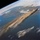 An orbital photograph taken from space showing Earth's surface with a large volcanic ash or dust plume extending diagonally across the frame over a body of water. The plume originates from a point source on a landmass in the upper right and disperses westward or southwestward across what appears to be an ocean or large sea. Snow-covered terrain, coastlines, and cloud formations are visible surrounding the plume.