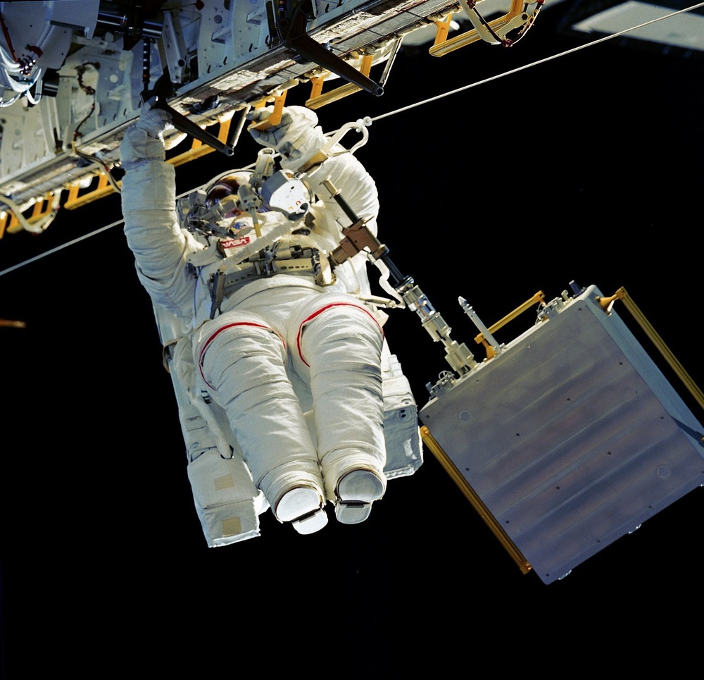 An astronaut in a white NASA EMU (Extravehicular Mobility Unit) spacesuit is performing a spacewalk (EVA) outside a spacecraft or space station. The astronaut is positioned horizontally relative to the camera, gripping a structural element of the spacecraft with one hand while working near equipment. The black void of space serves as the background, with spacecraft hardware visible in the upper portion of the frame.
