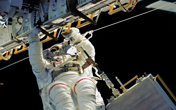 An astronaut in a white NASA EMU (Extravehicular Mobility Unit) spacesuit is performing a spacewalk (EVA) outside a spacecraft or space station. The astronaut is positioned horizontally relative to the camera, gripping a structural element of the spacecraft with one hand while working near equipment. The black void of space serves as the background, with spacecraft hardware visible in the upper portion of the frame.