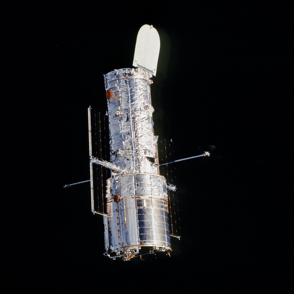 The image shows a large space telescope, consistent in appearance with the Hubble Space Telescope, photographed against the black void of space. The spacecraft is illuminated by sunlight on one side, revealing its cylindrical body wrapped in metallic thermal insulation blankets. Solar panel booms and antenna structures extend from the main body, and a light-colored aperture door is visible at the top.
