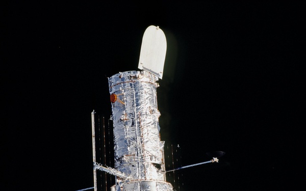 The image shows a large space telescope, consistent in appearance with the Hubble Space Telescope, photographed against the black void of space. The spacecraft is illuminated by sunlight on one side, revealing its cylindrical body wrapped in metallic thermal insulation blankets. Solar panel booms and antenna structures extend from the main body, and a light-colored aperture door is visible at the top.