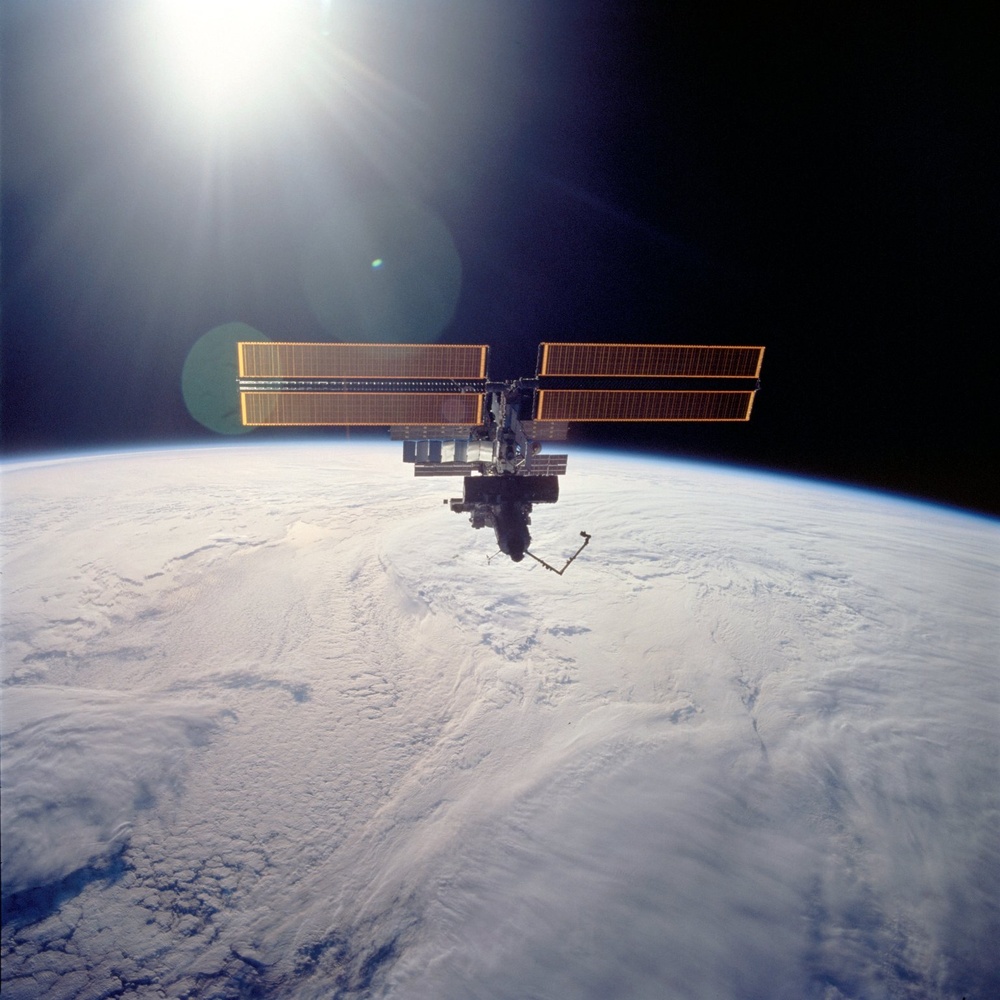 A space station is photographed in low Earth orbit against the blackness of space, with Earth's curved horizon and cloud-covered surface visible below. The station features large golden/orange solar panel arrays extending horizontally from a central module structure. The sun is visible in the upper left, causing lens flare artifacts in the image.