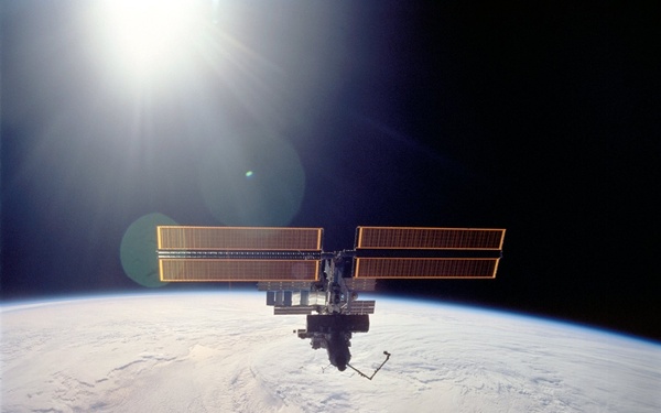 A space station is photographed in low Earth orbit against the blackness of space, with Earth's curved horizon and cloud-covered surface visible below. The station features large golden/orange solar panel arrays extending horizontally from a central module structure. The sun is visible in the upper left, causing lens flare artifacts in the image.