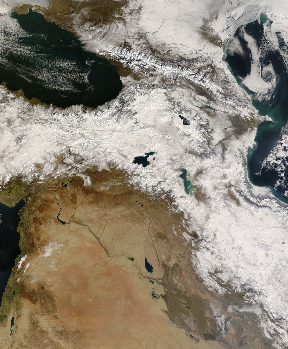 This is a satellite image of the Middle East and surrounding regions, showing a broad swath from the Eastern Mediterranean and Black Sea in the west to the Caspian Sea in the east. Extensive snow cover blankets the Anatolian Plateau, Caucasus Mountains, Zagros Mountains, and surrounding highland areas, contrasting sharply with the arid desert terrain of Syria, Iraq, and the Arabian Peninsula to the south. Several large water bodies are clearly visible, including the Black Sea, Caspian Sea, and inland lakes.