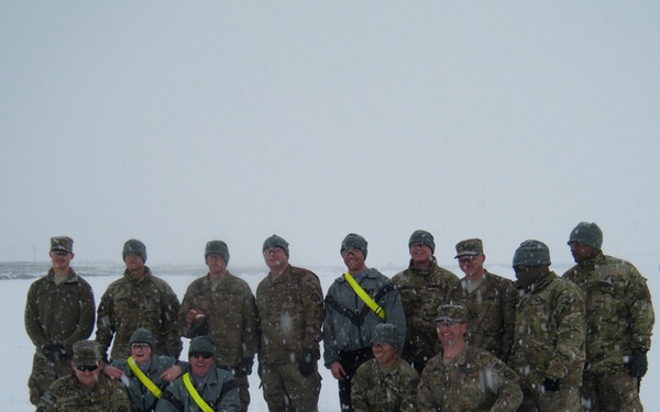 A group of approximately 15 military personnel posing for a group photo in heavy snowfall conditions. They are wearing camouflage uniforms with cold-weather gear including wool caps and gloves. Several individuals are wearing high-visibility yellow/neon reflective sashes or belts.