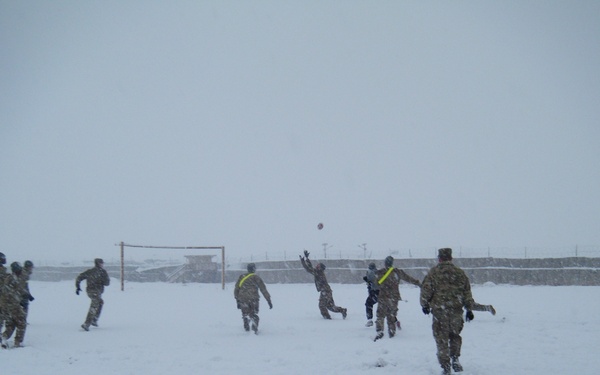 A group of military personnel in camouflage uniforms are playing an outdoor ball sport, likely volleyball or football, on a snow-covered field during heavy snowfall. Some players are wearing yellow/neon safety vests to distinguish teams. A goal post is visible in the background, along with what appears to be a military installation perimeter wall.