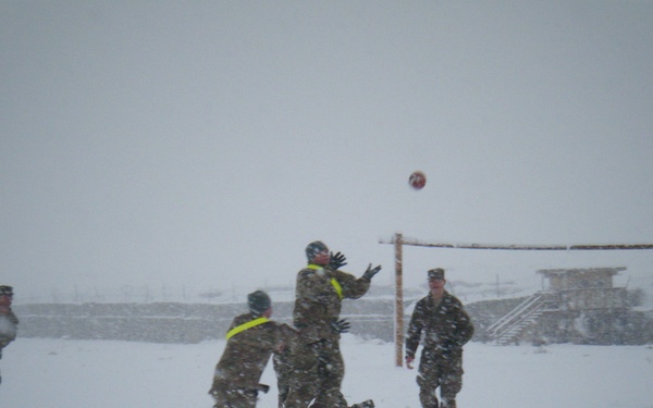 A group of military personnel in camouflage uniforms are playing soccer on a snow-covered field during a heavy snowstorm. A red ball is visible in the air near a makeshift goal post, with players actively engaged in the game. The setting appears to be a forward operating base or military installation, visible in the background.
