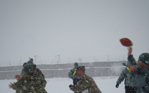 A group of military personnel in camouflage uniforms are playing American football in heavy snowfall conditions. The game appears to be taking place within a military installation, visible by the perimeter wall and security infrastructure in the background. Despite the harsh winter weather, the participants appear actively engaged in the recreational activity.