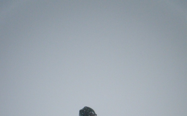 A single person stands in an open, snow-covered field during an active snowstorm with low visibility. The individual is dressed in full camouflage winter gear and appears to be holding a small object, possibly a radio or cigar, to their face. The overcast, whiteout conditions and blowing snow dominate the scene.