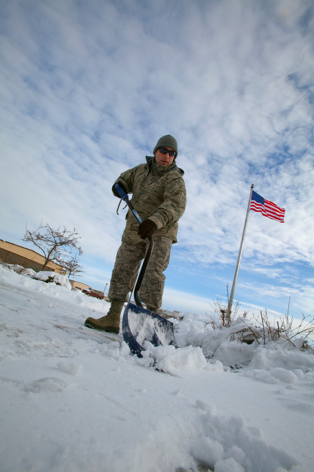 A military service member in camouflage uniform is shoveling snow from a pathway or walkway at what appears to be a military installation. The photo is taken from a low angle, giving a dramatic perspective with the subject dominating the foreground. An American flag on a flagpole is visible in the background against a partly cloudy sky.