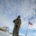 108th Wing removes snow from winter storm Nemo A military service member in camouflage uniform is shoveling snow from a pathway or walkway at what appears to be a military installation. The photo is taken from a low angle, giving a dramatic perspective with the subject dominating the foreground. An American flag on a flagpole is visible in the background against a partly cloudy sky.
