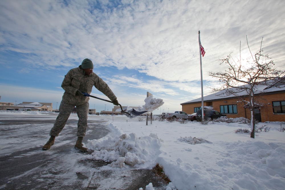 A military service member in camouflage uniform is shoveling snow from a paved area, likely at a military installation. An American flag flies at half or full staff on a flagpole in the background, with a brick building and snow-covered grounds visible. The scene depicts winter maintenance duties at what appears to be a U.S. military base.