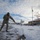 108th Wing removes snow from winter storm Nemo A military service member in camouflage uniform is shoveling snow from a paved area, likely at a military installation. An American flag flies at half or full staff on a flagpole in the background, with a brick building and snow-covered grounds visible. The scene depicts winter maintenance duties at what appears to be a U.S. military base.