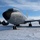 108th Wing removes snow from winter storm Nemo A U.S. Air Force KC-135 Stratotanker aircraft (serial number 3544) assigned to the 108th Wing, New Jersey Air National Guard, is parked on a snow-covered tarmac. The aircraft is photographed from a low front-quarter angle under clear blue skies with light cloud cover. The engine nacelles display distinctive orange '108 Wing / New Jersey ANG' markings with a tiger emblem.