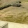 An aerial or elevated view of what appears to be a military or law enforcement training range. A V-shaped or angled barrier constructed from stacked rubber tires is visible, with black smoke rising from a burning section near the apex. A single figure in tactical gear is visible in the lower-left corner behind a concrete structure.