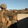 Two military personnel in MARPAT (Marine pattern) camouflage uniforms and tactical gear are positioned behind a concrete block wall in what appears to be a training exercise. One individual is pointing or gesturing with an outstretched arm, likely indicating a direction or target. The setting appears to be a military training facility with hilly terrain visible in the background.