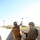 Two U.S. Marines in MARPAT digital camouflage uniforms and tactical gear are conducting a grenade throwing exercise at what appears to be a military training range. One Marine, identified by name tape as 'WALKER,' is in the act of throwing a grenade over a concrete block wall while the second Marine observes and assists. The setting is an outdoor training facility with arid terrain visible in the background.
