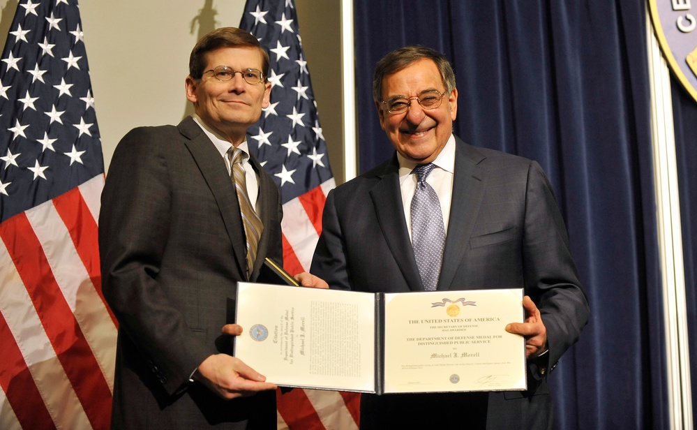 Two men in business suits are posing together while holding an open certificate or award document. The document appears to be a Department of Defense Medal for Distinguished Public Service awarded to 'Michael J. Morell.' The setting includes American flags and an official seal, suggesting a formal government ceremony.