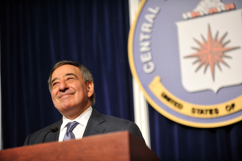 A man in a dark suit and patterned tie stands at a wooden podium, smiling upward. Behind him is a large official seal displaying the words 'CENTRAL' and 'UNITED STATES OF,' consistent with the CIA seal, mounted against a dark blue draped curtain backdrop. The setting appears to be a formal press conference or official address.