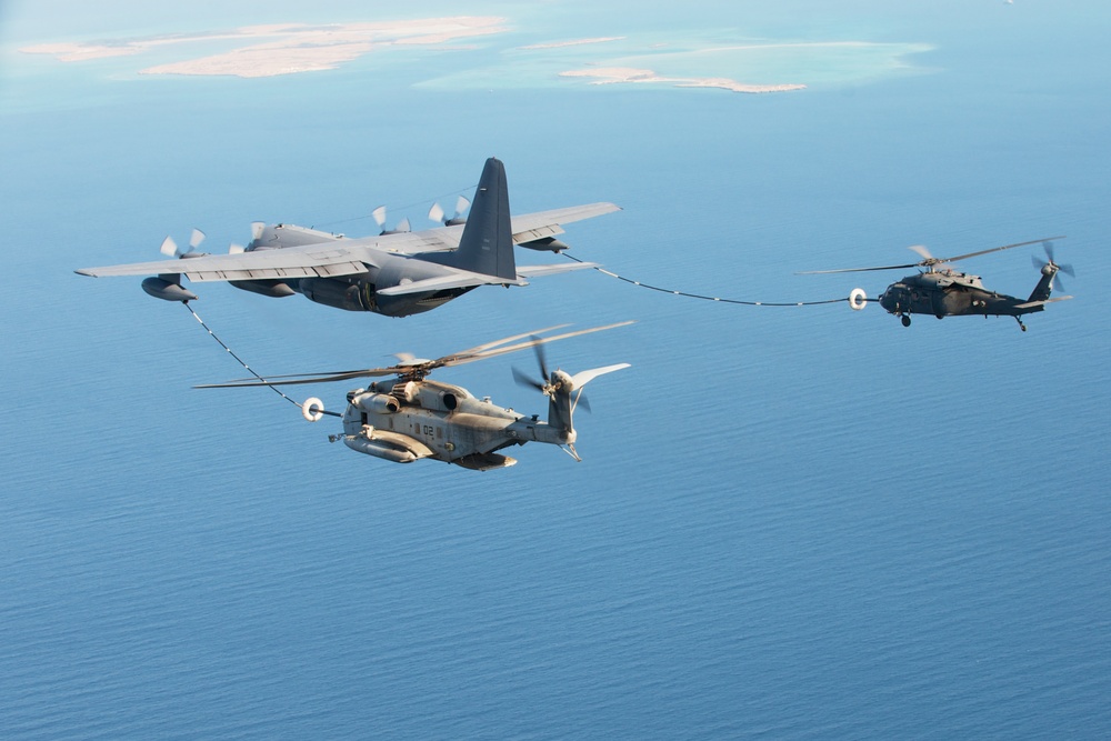 An aerial refueling operation is taking place over a body of water, with a fixed-wing tanker aircraft simultaneously refueling two military helicopters via extended fuel hoses. The tanker aircraft appears to be a C-130 variant configured for aerial refueling, transferring fuel to a large heavy-lift helicopter on the left and a medium utility helicopter on the right. A coastline with sandy terrain is visible in the background.