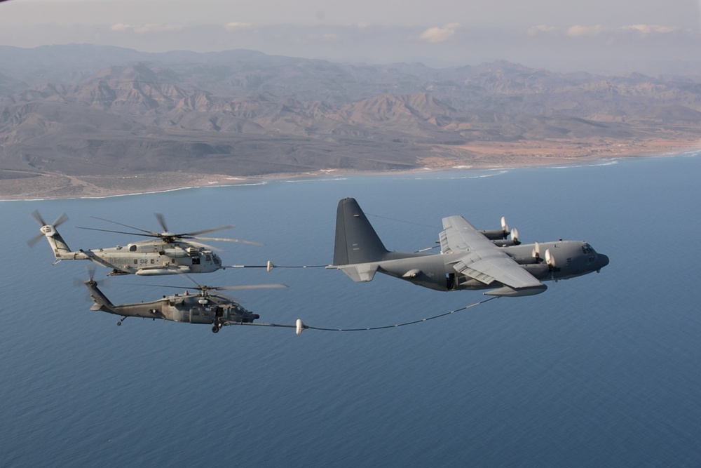 An aerial refueling operation is underway over a coastal region with arid mountainous terrain visible in the background. A C-130 Hercules tanker aircraft is simultaneously refueling two military helicopters — one larger and one smaller — using hose-and-drogue systems extended from both wings. The aircraft appear to be U.S. military assets operating over what appears to be a desert coastline.
