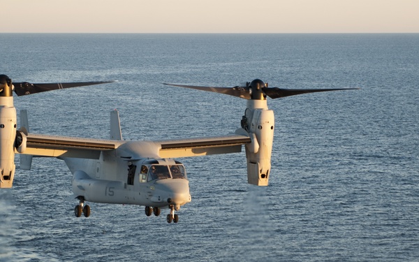 A military tiltrotor aircraft, identified as a Bell Boeing V-22 Osprey, is hovering or approaching to land on the deck of a naval vessel over open ocean. The aircraft bears the number '15' on its nose and appears to be in helicopter mode with rotors tilted upward. The image was taken during golden hour lighting conditions, suggesting early morning or late afternoon.
