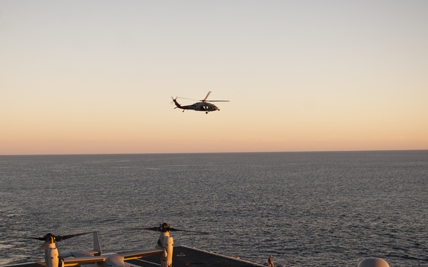 A military naval vessel's flight deck is captured at sunset or sunrise over open ocean. An MV-22 Osprey tiltrotor aircraft bearing the number '15' is parked on the deck while a helicopter, consistent with an MH-60 Sikorsky, hovers overhead. Flight deck crew in yellow and white jerseys are visible conducting flight operations.