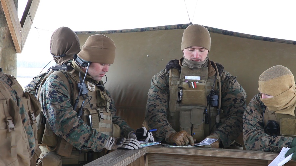 Three military personnel in MARPAT (Marine pattern) camouflage uniforms are gathered around a wooden field table, reviewing documents or maps in what appears to be a forward operating position or field command post. The soldiers are equipped with tactical gear including body armor, communications equipment, and cold-weather headgear, suggesting operations in a cold environment. The setting appears to be a covered outdoor structure used as a temporary command or briefing station.