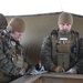 Three military personnel in MARPAT (Marine pattern) camouflage uniforms are gathered around a wooden field table, reviewing documents or maps in what appears to be a forward operating position or field command post. The soldiers are equipped with tactical gear including body armor, communications equipment, and cold-weather headgear, suggesting operations in a cold environment. The setting appears to be a covered outdoor structure used as a temporary command or briefing station.