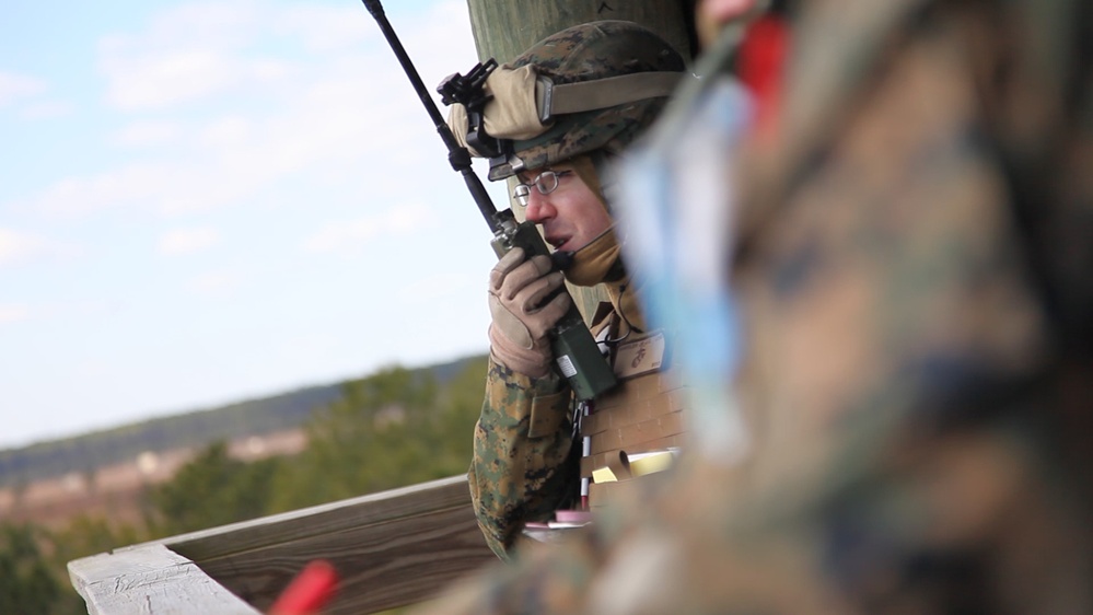 A military service member in full combat gear, including a camouflage helmet and body armor, is speaking into a handheld radio while positioned behind a wooden structure. The individual is wearing glasses and tactical gloves, suggesting a communications or forward observer role. A second service member in camouflage is partially visible and out of focus in the foreground.