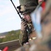 A military service member in full combat gear, including a camouflage helmet and body armor, is speaking into a handheld radio while positioned behind a wooden structure. The individual is wearing glasses and tactical gloves, suggesting a communications or forward observer role. A second service member in camouflage is partially visible and out of focus in the foreground.