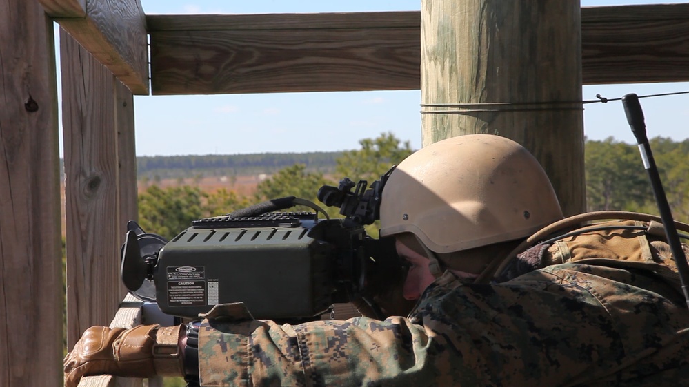 A military service member in MARPAT camouflage uniform and tan helmet is aiming a weapon system from an elevated wooden platform or tower. The weapon appears to be a crew-served or specialized system with a boxy receiver and optics mounted on top. The setting appears to be a training range with open terrain and tree lines visible in the background.