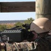 A military service member in MARPAT camouflage uniform and tan helmet is aiming a weapon system from an elevated wooden platform or tower. The weapon appears to be a crew-served or specialized system with a boxy receiver and optics mounted on top. The setting appears to be a training range with open terrain and tree lines visible in the background.