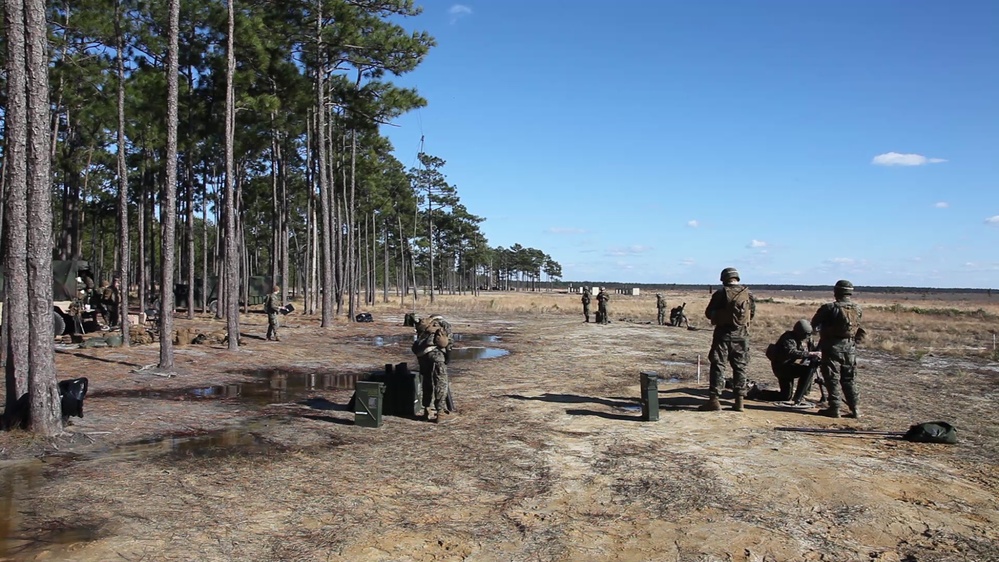 The image shows military personnel in camouflage uniforms conducting what appears to be a live-fire range exercise at an outdoor training facility. The setting is a flat, open sandy range bordered by tall pine trees on the left side, consistent with training installations in the southeastern United States. Multiple personnel are positioned at firing lines with ammunition cans visible on the ground.