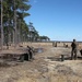 The image shows military personnel in camouflage uniforms conducting what appears to be a live-fire range exercise at an outdoor training facility. The setting is a flat, open sandy range bordered by tall pine trees on the left side, consistent with training installations in the southeastern United States. Multiple personnel are positioned at firing lines with ammunition cans visible on the ground.
