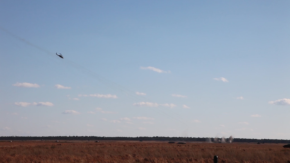 An open, dry grassland or field under a partly cloudy blue sky, with a helicopter visible in the upper left portion of the image. On the ground in the mid-right area, there are visible dust or smoke plumes suggesting recent impacts or explosions. The scene appears consistent with a military training exercise or live-fire demonstration.