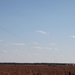 An open, dry grassland or field under a partly cloudy blue sky, with a helicopter visible in the upper left portion of the image. On the ground in the mid-right area, there are visible dust or smoke plumes suggesting recent impacts or explosions. The scene appears consistent with a military training exercise or live-fire demonstration.