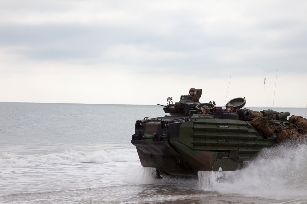 An amphibious assault vehicle (AAV), likely a US Marine Corps AAV-7A1, is emerging from the surf onto a beach during what appears to be an amphibious landing exercise. The vehicle is painted in green and brown camouflage and carries personnel and equipment on its exterior. The overcast sky and churning water suggest active coastal operations.