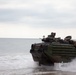 An amphibious assault vehicle (AAV), likely a US Marine Corps AAV-7A1, is emerging from the surf onto a beach during what appears to be an amphibious landing exercise. The vehicle is painted in green and brown camouflage and carries personnel and equipment on its exterior. The overcast sky and churning water suggest active coastal operations.