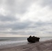 Two amphibious assault vehicles (AAVs) are shown on a sandy beach, having emerged from or moving along the shoreline under an overcast sky. The vehicles appear to be military tracked amphibious personnel carriers, consistent with U.S. Marine Corps AAV7A1 models, with personnel visible on top. A vessel is faintly visible on the horizon, suggesting a naval operation or amphibious landing exercise.