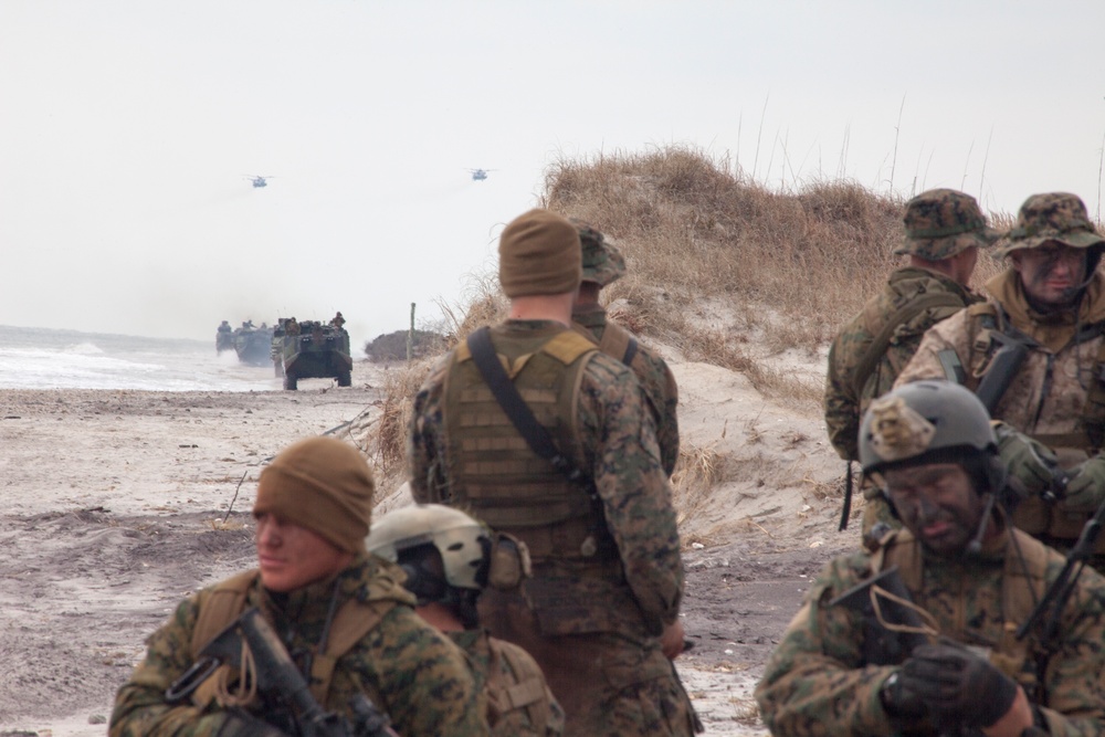 The image depicts a military amphibious assault exercise on a beach, with Marines in MARPAT camouflage uniforms taking positions along sand dunes. Amphibious Assault Vehicles (AAVs) are visible emerging from the surf zone, and at least two helicopters can be seen in the hazy background sky. The scene is consistent with a large-scale amphibious landing drill, likely conducted by the United States Marine Corps.