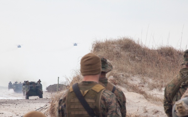 The image depicts a military amphibious assault exercise on a beach, with Marines in MARPAT camouflage uniforms taking positions along sand dunes. Amphibious Assault Vehicles (AAVs) are visible emerging from the surf zone, and at least two helicopters can be seen in the hazy background sky. The scene is consistent with a large-scale amphibious landing drill, likely conducted by the United States Marine Corps.