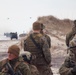 The image depicts a military amphibious assault exercise on a beach, with Marines in MARPAT camouflage uniforms taking positions along sand dunes. Amphibious Assault Vehicles (AAVs) are visible emerging from the surf zone, and at least two helicopters can be seen in the hazy background sky. The scene is consistent with a large-scale amphibious landing drill, likely conducted by the United States Marine Corps.